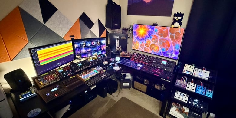 Music studio with three screens, keyboards and audio equipment beneath geometric wall panels.